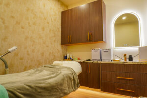 Interior view of a modern treatment room featuring a massage table draped with a soft blanket, wooden cabinetry with gold handles, and a well-lit mirror. The space is designed for relaxation, equipped with beauty products and tools, reflecting a serene atmosphere.